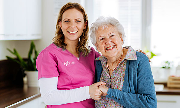 Happy senior woman standing with a female AgeSMART caregiver wearing a pink shirt, representing compassionate non-medical in-home senior care and support